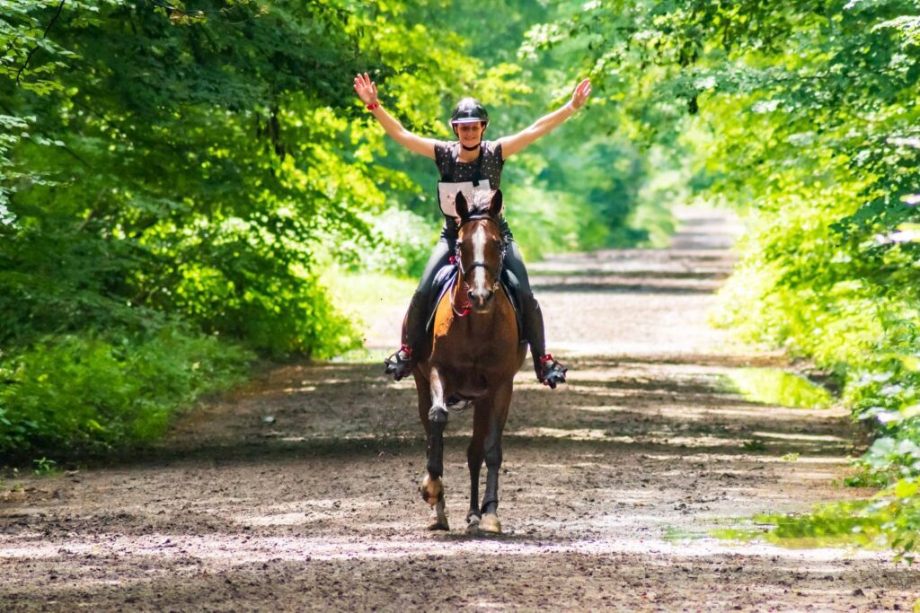 Cheval au galop dans une forêt, la cavalière lève les deux bras